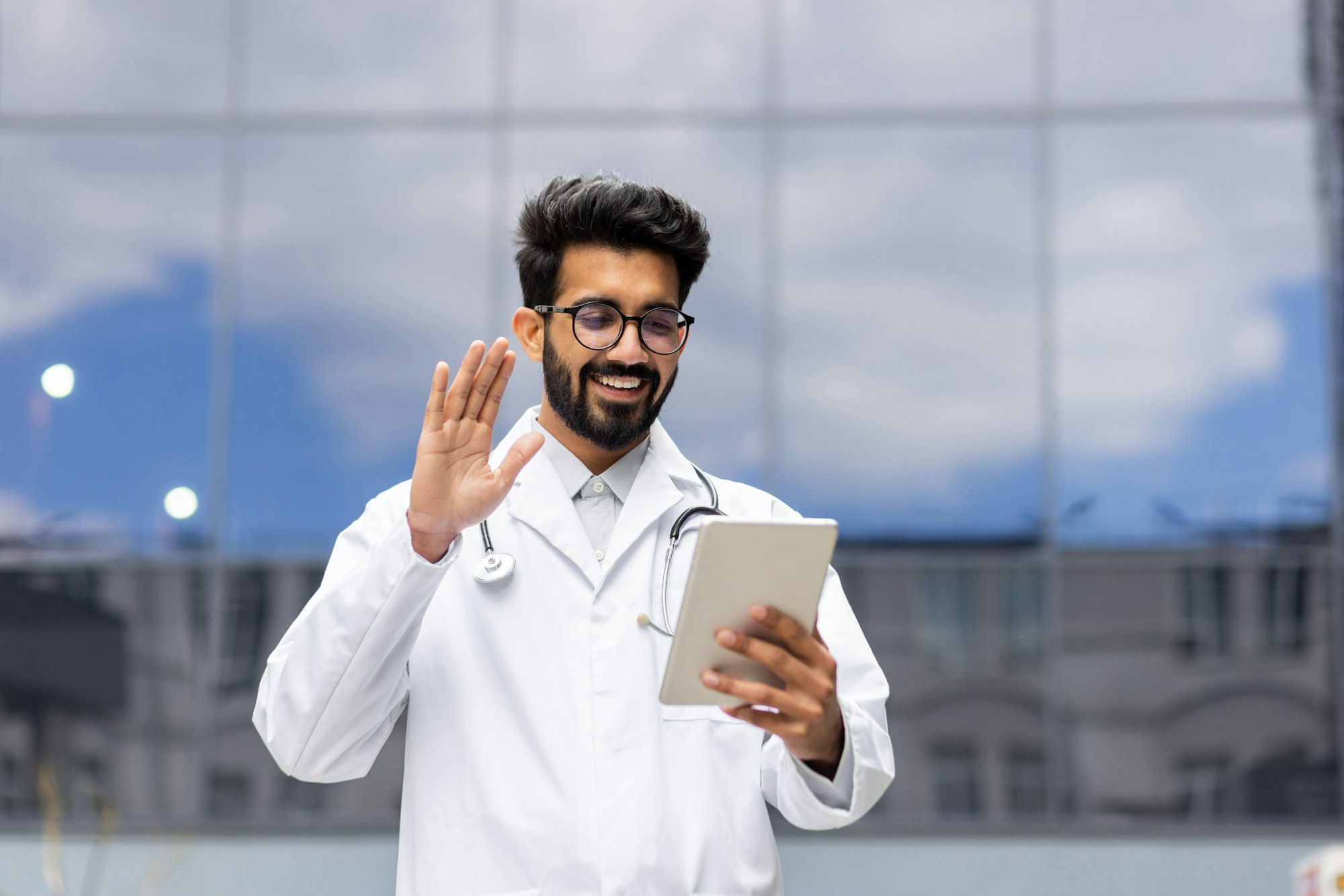 Young male doctor in white medical coat outdoors, smiling and talking consulting patients remotely